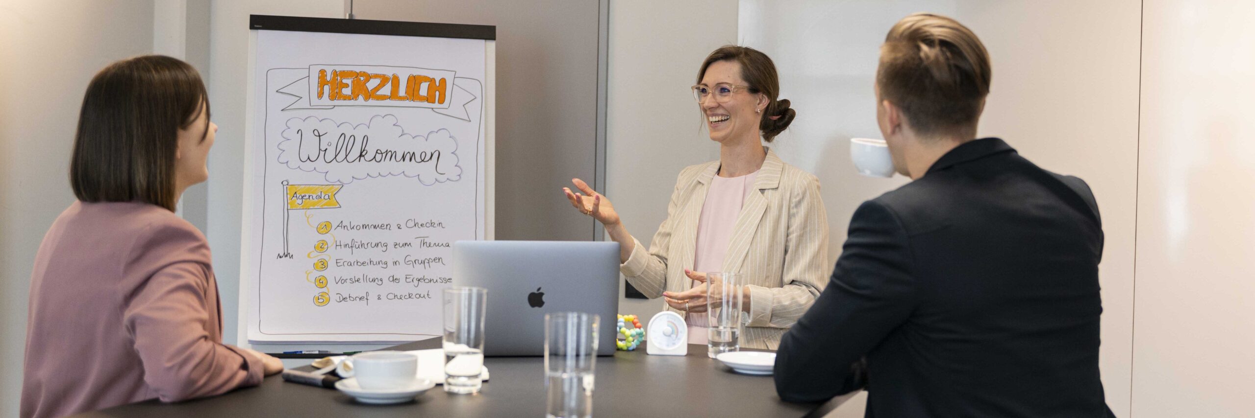 JanineFrank-Projektmanagement-Kundenworshop Janine Frank steht erklärend vor einem Flipchart. Am Tisch vor ihr sitzen zwei Kunde mit dem Rücken zur Kamera und hören aufmerksam zu.